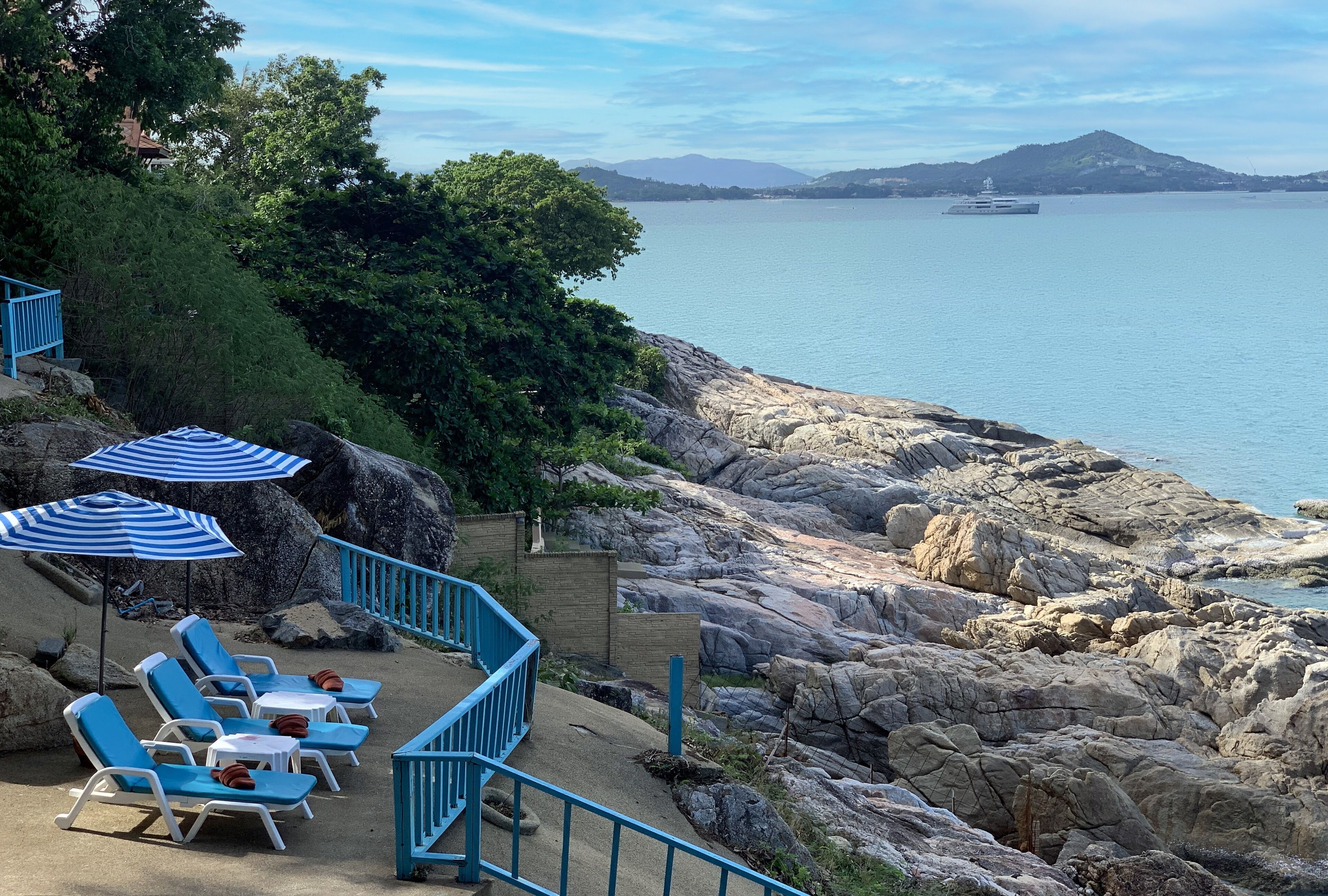 Eine Küsten-Terrasse mit blauen Liegestühlen und Sonnenschirmen, mit Blick auf eine felsige Küstenlinie und ruhiges Meer.