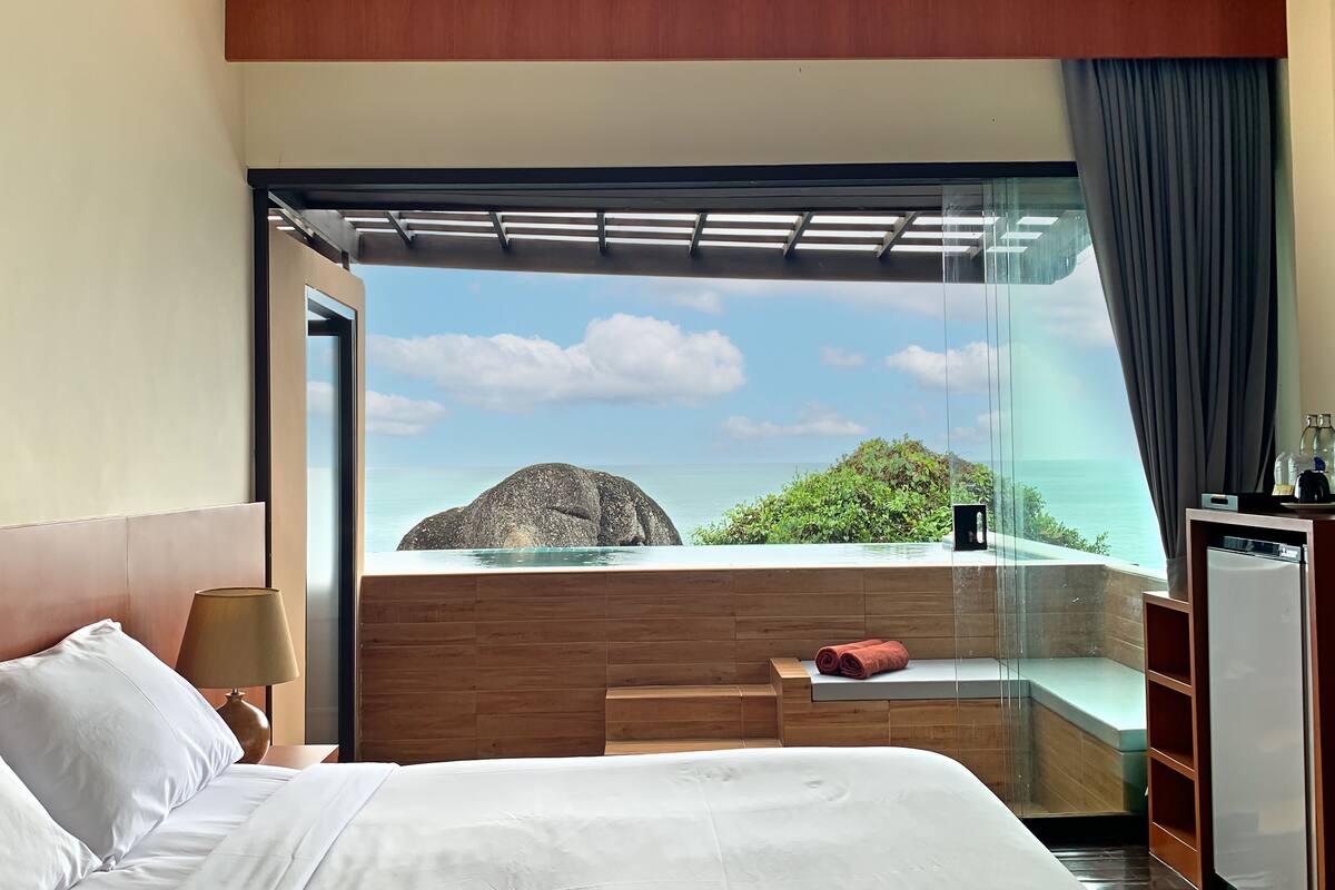 Habitación de hotel con cama, mesita de noche, lámpara, banca de madera y vista al océano y una roca grande.