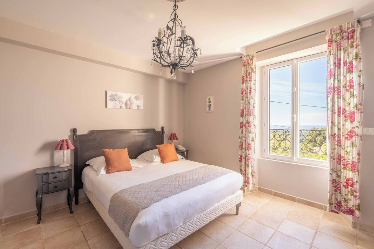 Une chambre à coucher avec un lit, des tables de chevet, un lustre et une fenêtre avec des rideaux.