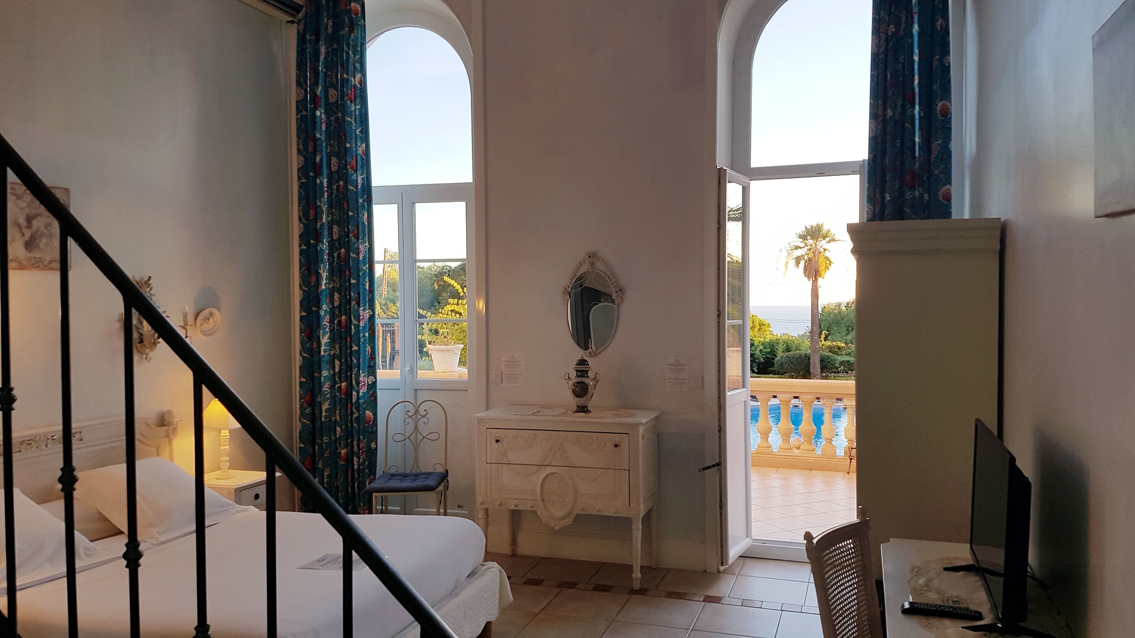 A bedroom with a bed, a dresser, a mirror, and a desk. There is a balcony with a view of the sea and palm trees.