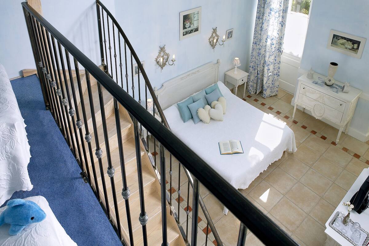 Une chambre à coucher comprenant un lit, une table de chevet, une commode, une fenêtre avec des rideaux et un escalier.