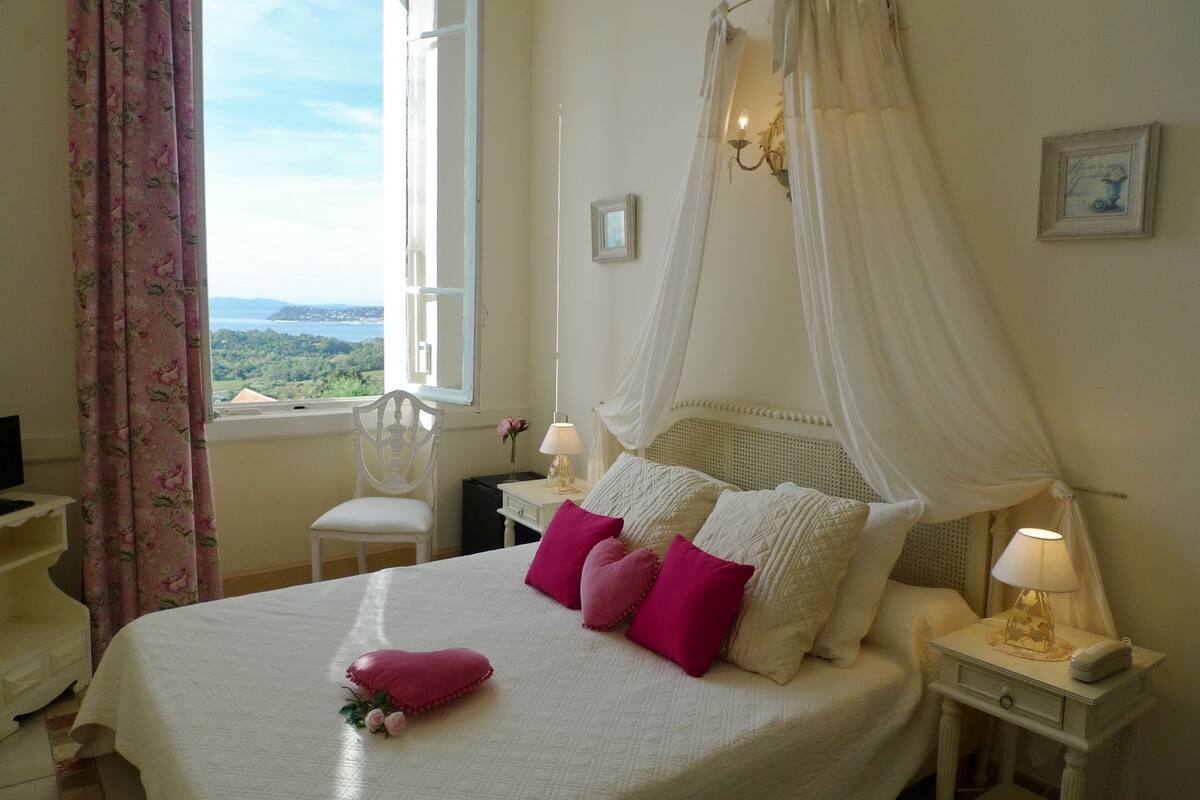 Une chambre à coucher comprenant un lit, une table de chevet, une chaise, une fenêtre donnant sur un beau paysage et une petite table placée près du lit avec une lampe.