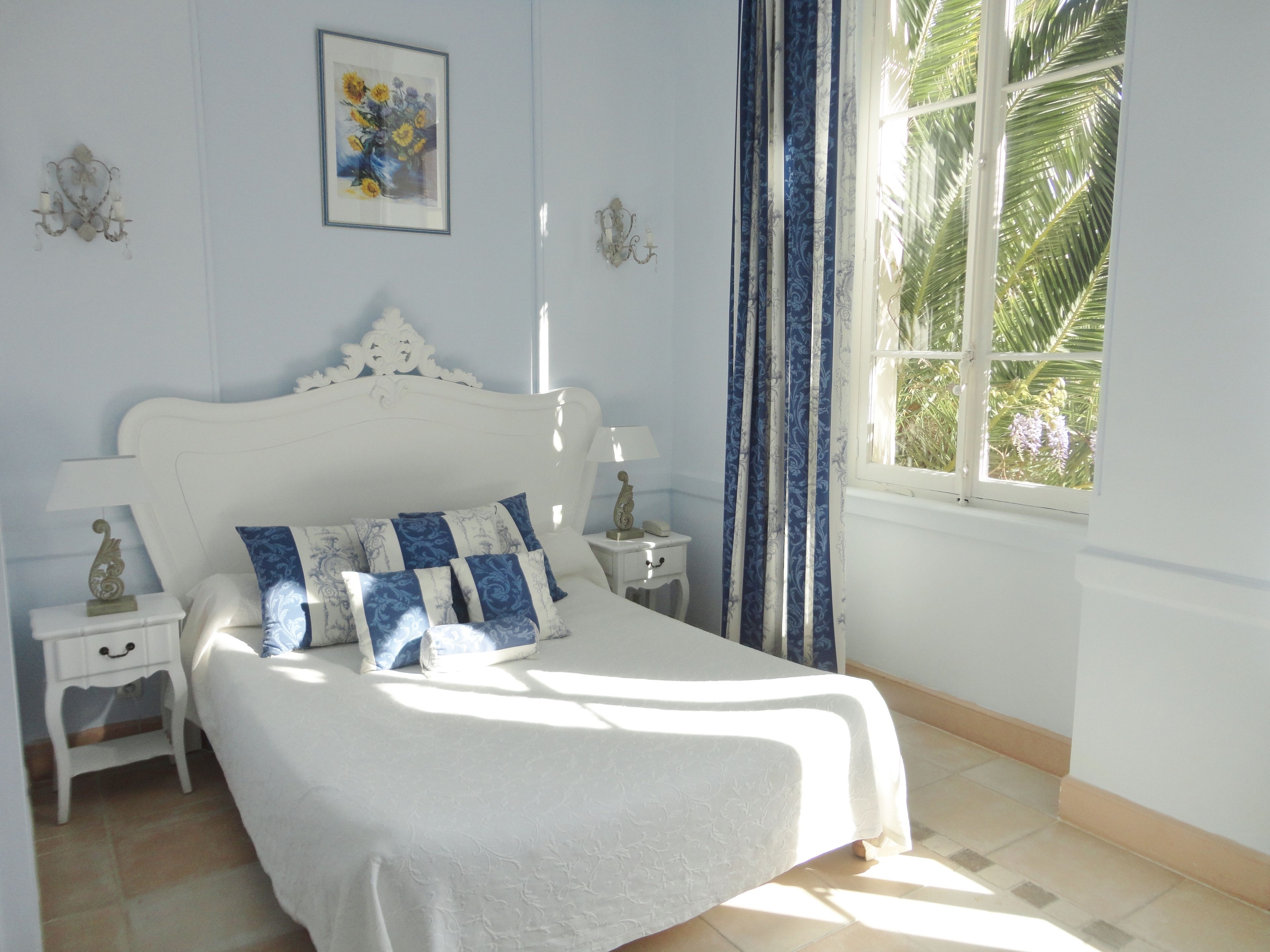Un dormitorio con una cama blanca, dos mesitas de noche, un velador, una ventana con cortinas y un aplique de pared.