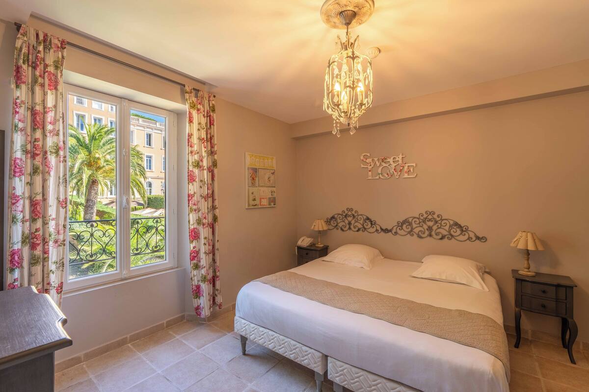 Une chambre à coucher avec un grand lit, un lustre, une table de chevet, un bureau et une fenêtre avec des rideaux.
