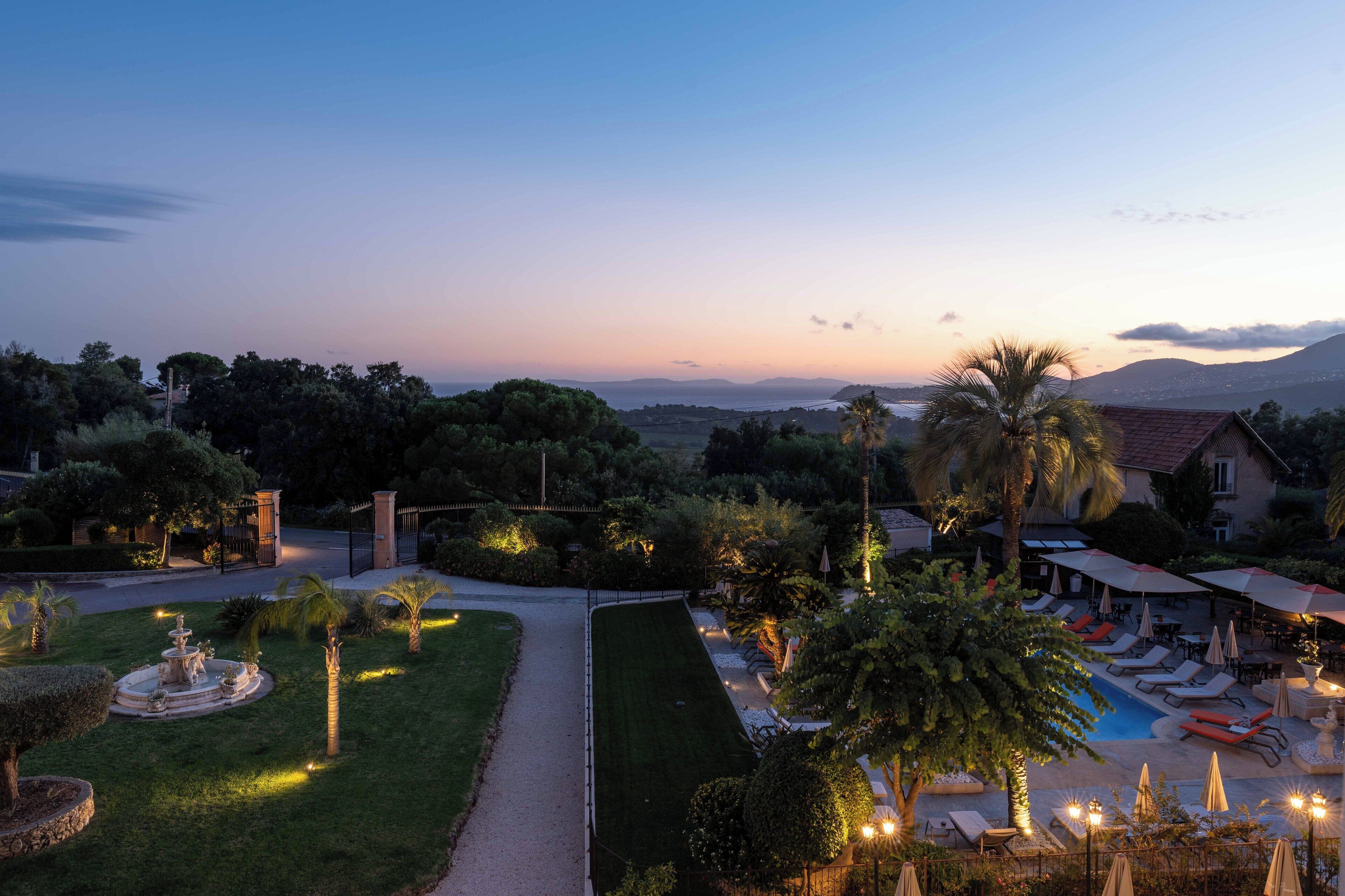 A landscaped garden with a fountain, a swimming pool with lounge chairs, and a view of the sea and mountains.