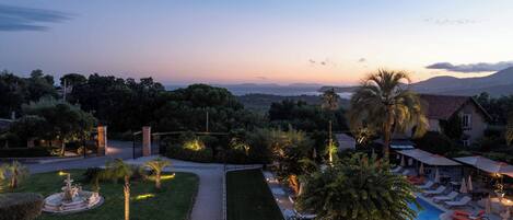 A landscaped garden with a fountain, a swimming pool with lounge chairs, and a view of the sea and mountains.