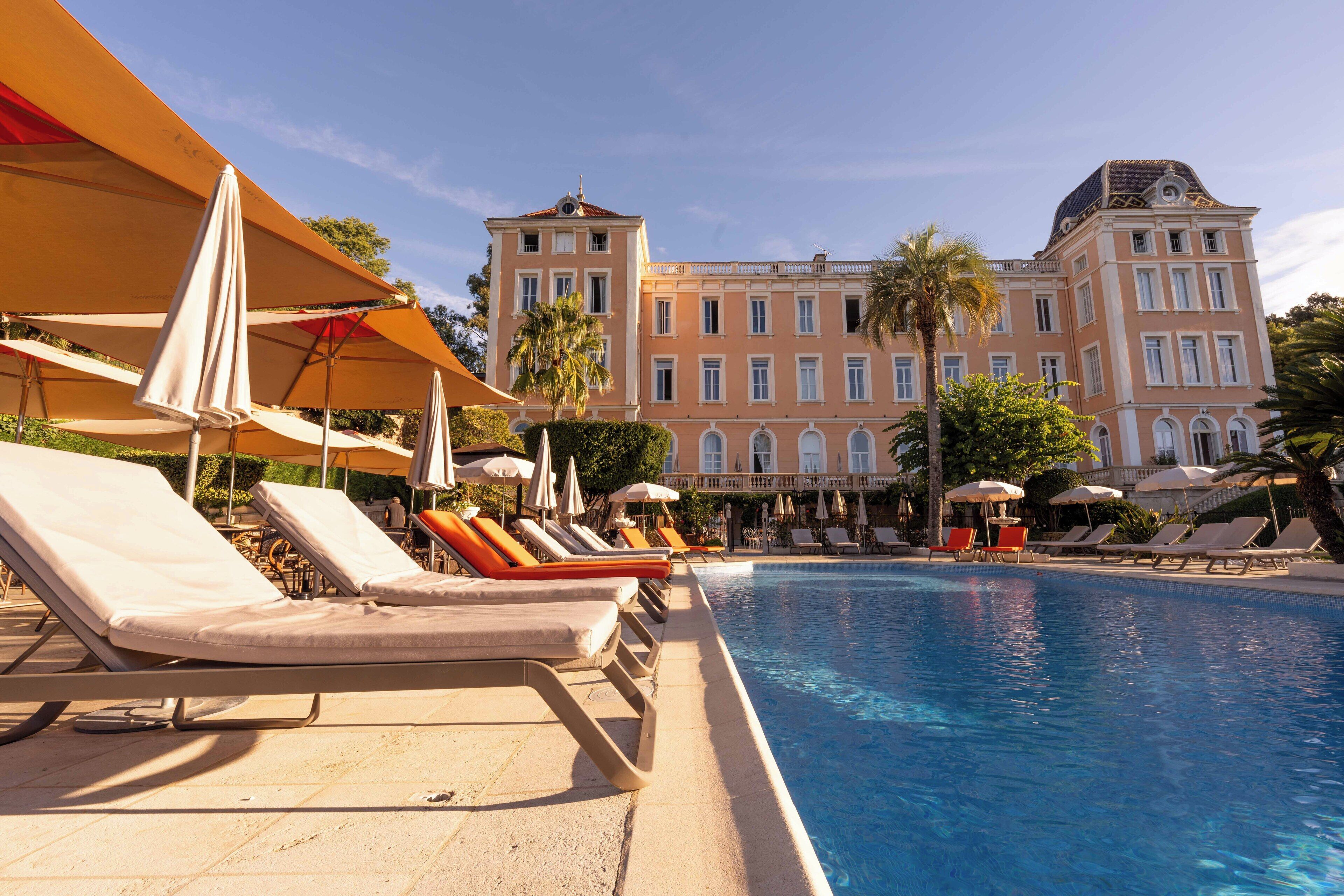 A poolside area with lounge chairs and umbrellas, a large building with classical architecture in the background, and a clear blue sky.