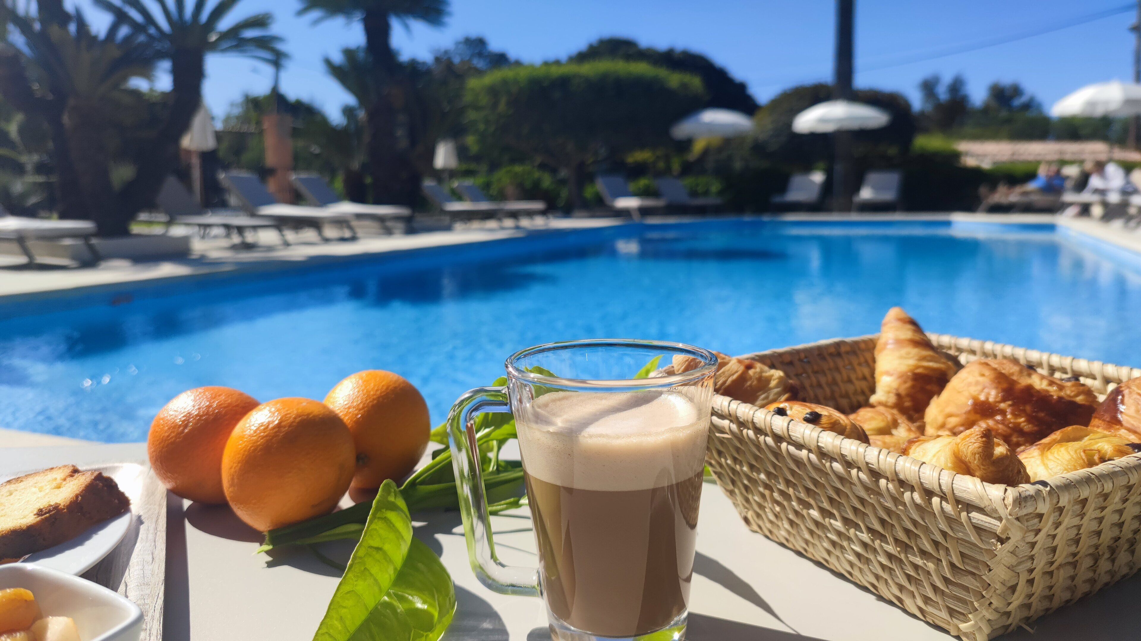 A poolside setting with a glass of iced coffee, oranges, bread, and a basket of croissants.