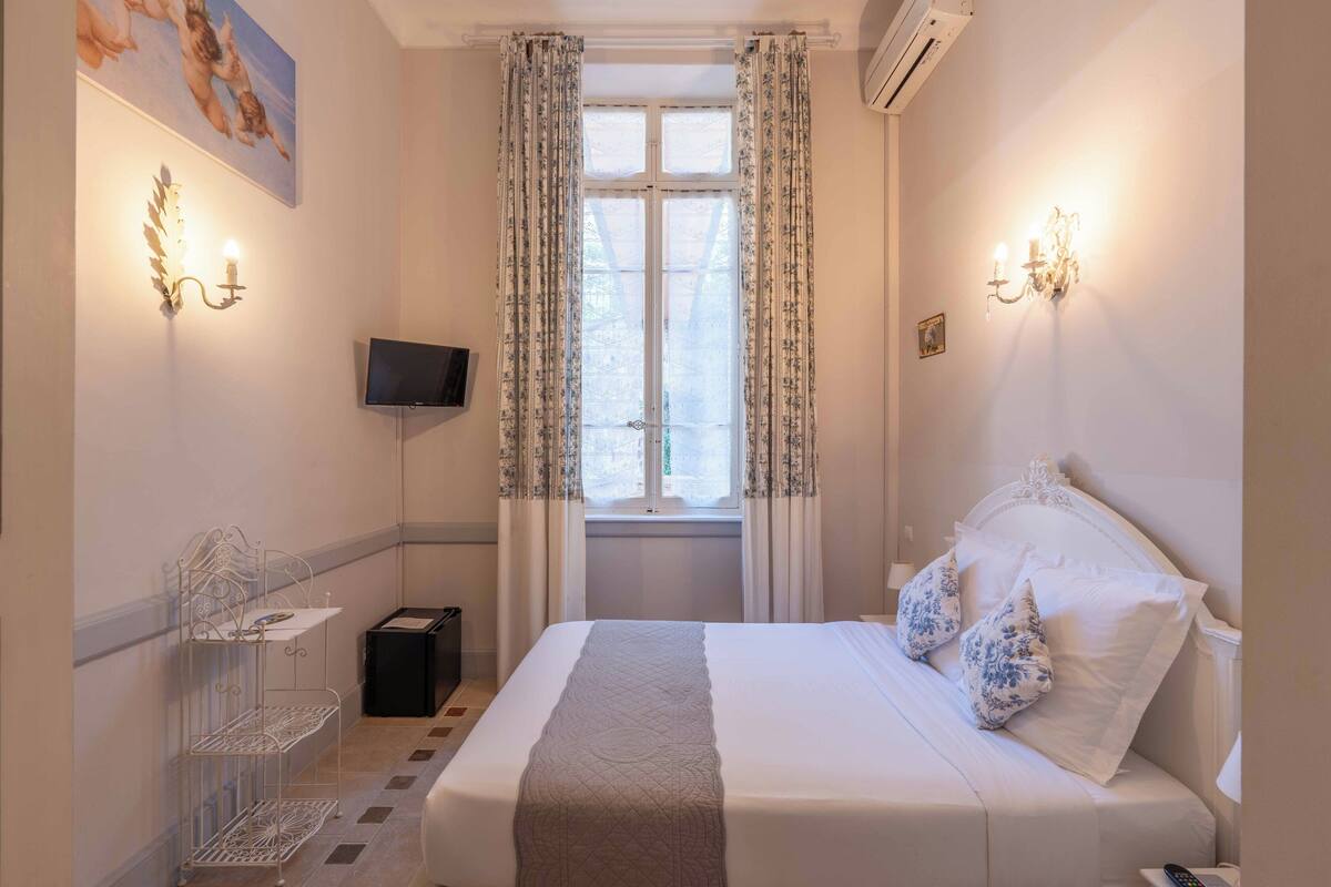 Une chambre d’hôtel équipée d’un lit, d’une télévision, d’une fenêtre avec des rideaux, d’une lampe fixée au mur et d’une étagère sur laquelle se trouve un téléviseur à écran plat.