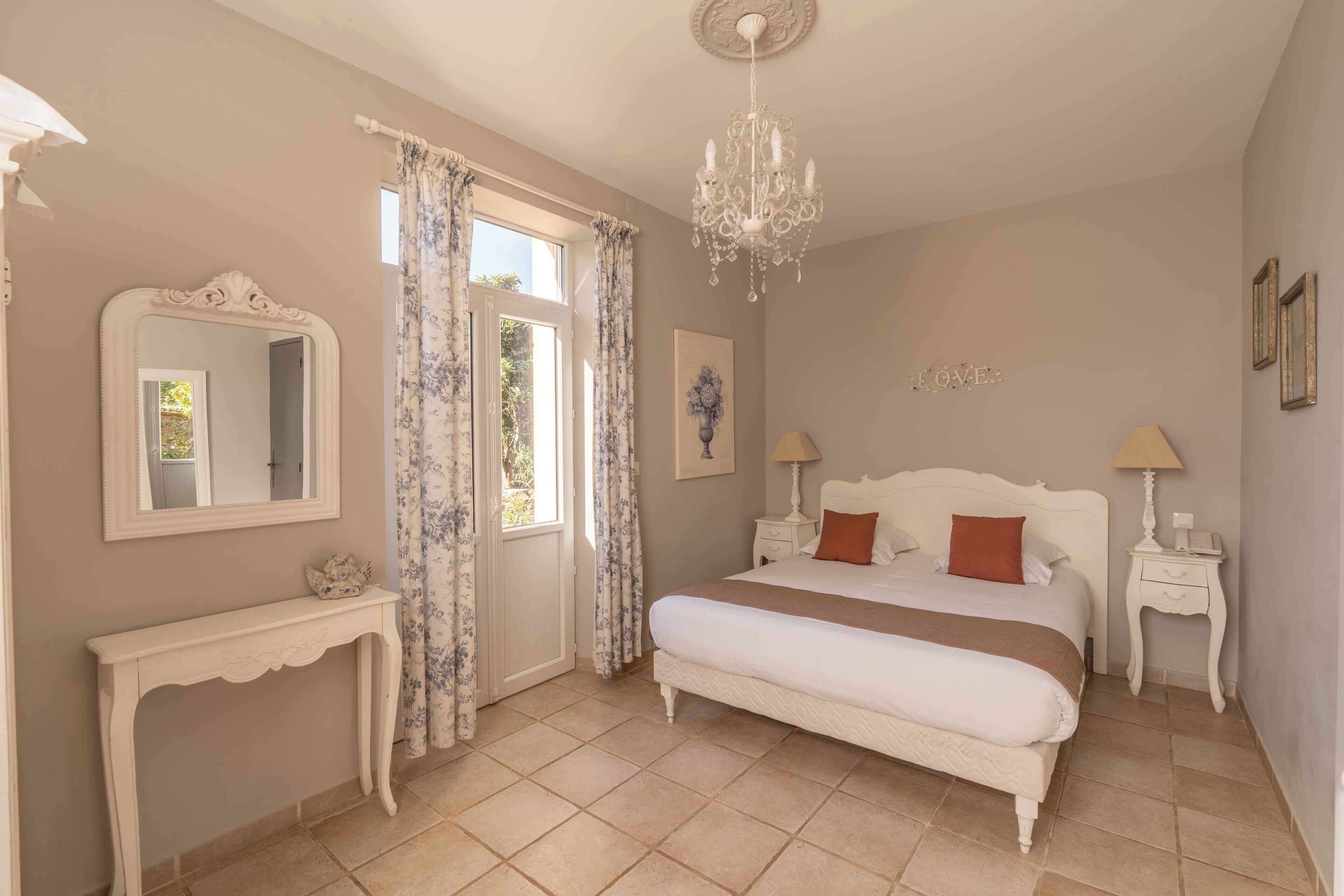 A bedroom with a large bed, a dresser, a mirror, a chandelier, and a window with curtains.