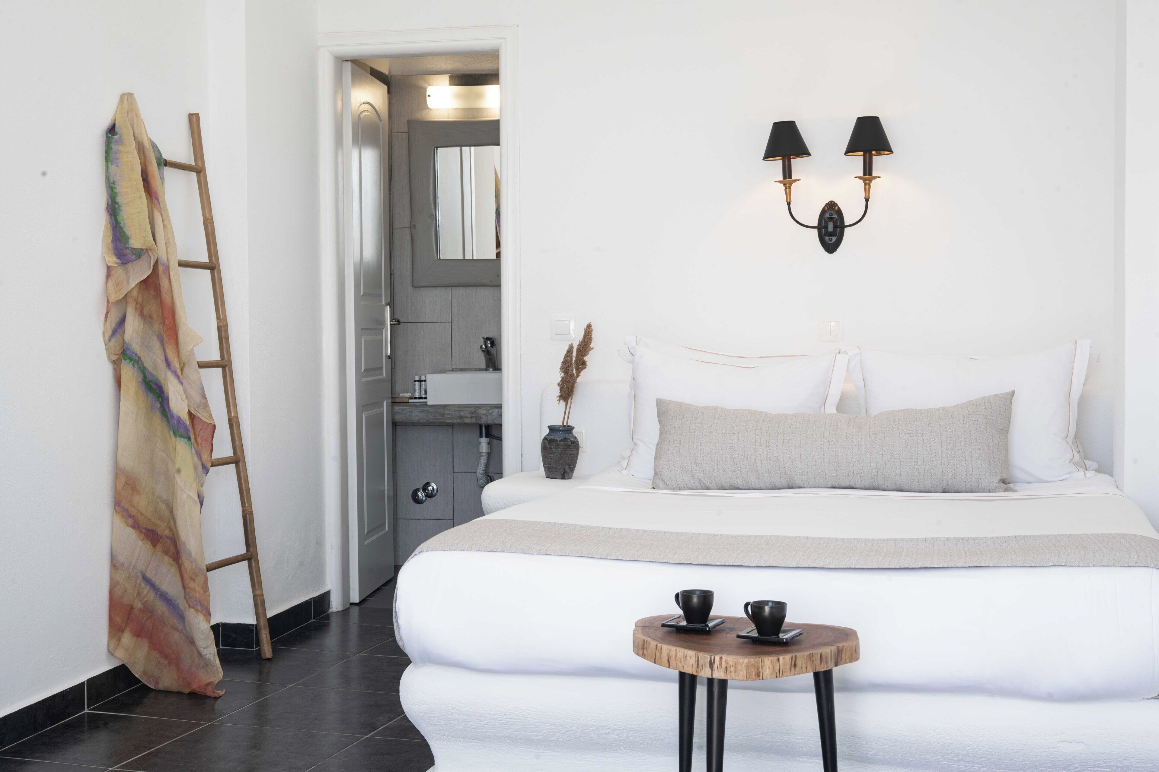 Un dormitorio moderno con una cama grande, una mesita redonda pequeña con dos tazas, una escalera apoyada en la pared y un baño visible a través de una puerta abierta.