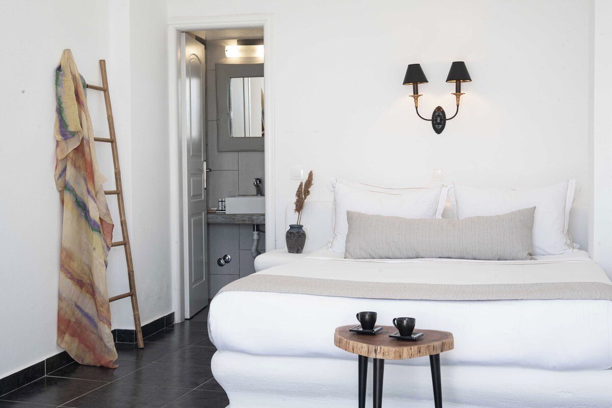 A modern bedroom with a large bed, a small round table with two cups, a ladder leaning against the wall, and a bathroom visible through an open door.