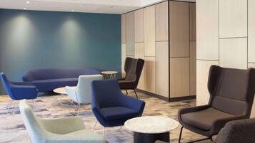 Un área de descanso moderna con un sofá azul, varios sillones, una mesita redonda y un panel divisor de madera.