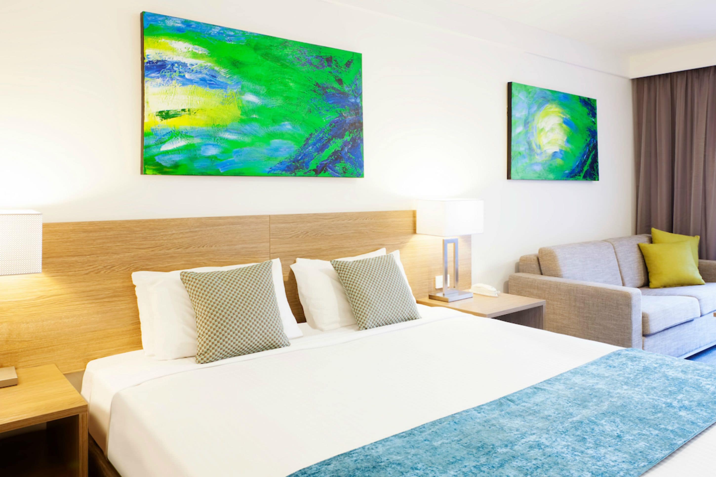 Una habitació d'hotel amb un llit gran, un panell de fusta al capçal, dos quadres a la paret, un sofà amb un coixí groc i una catifa blava decorativa.