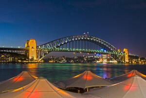 A large, illuminated bridge with two towers, over a body of water, with tents in the foreground.