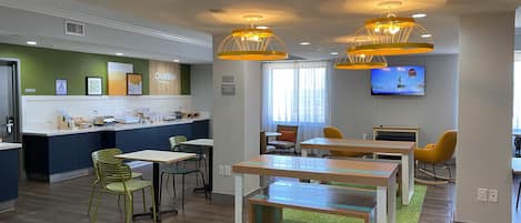 Un hall d’hôtel moderne doté d’un buffet pour le petit-déjeuner, d’un espace salon et d’un téléviseur diffusant une chaîne d’information.