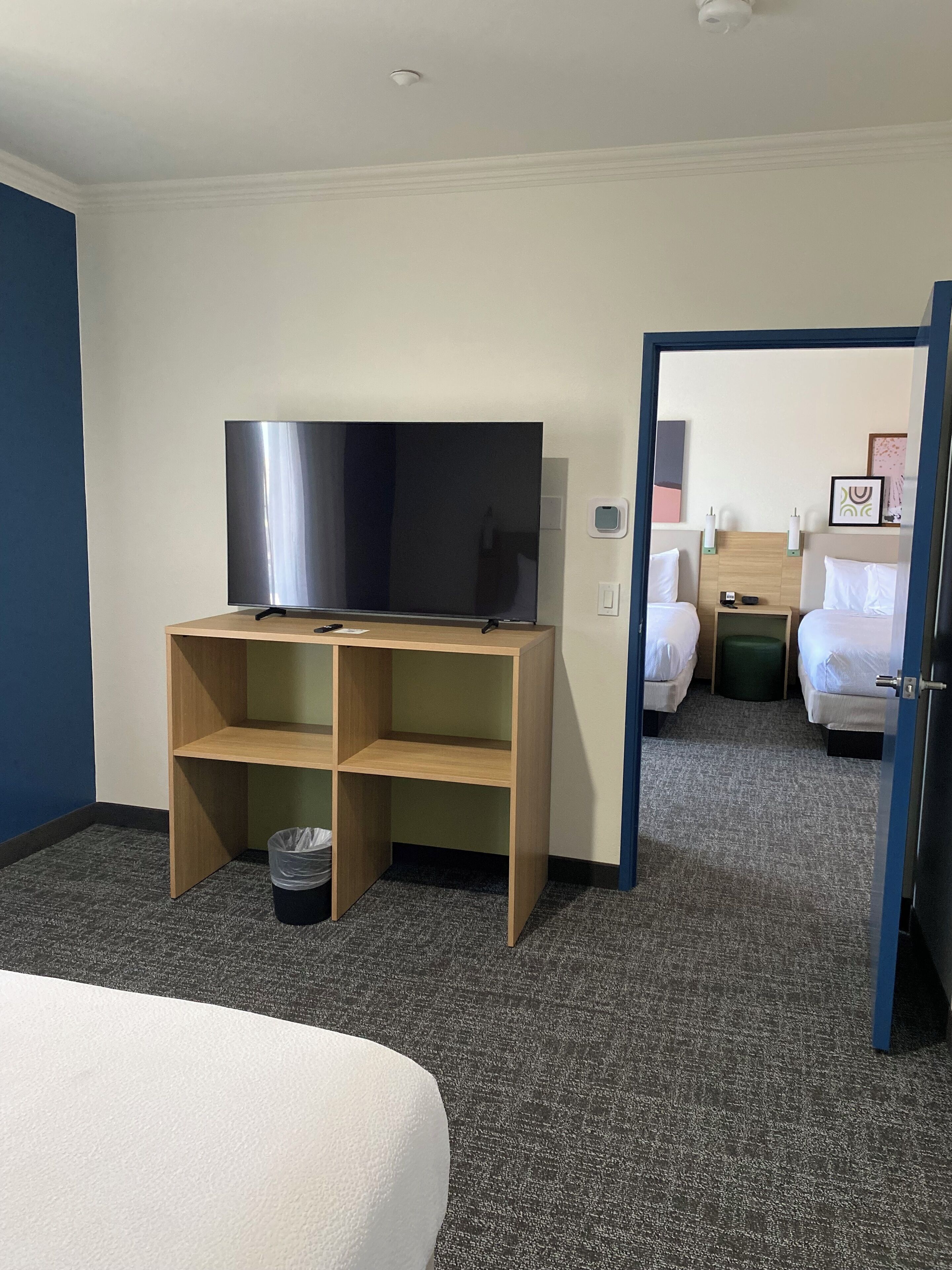A hotel room with a flat-screen TV on a wooden stand, a bed with white linens, a small trash bin, and a door leading to another room.