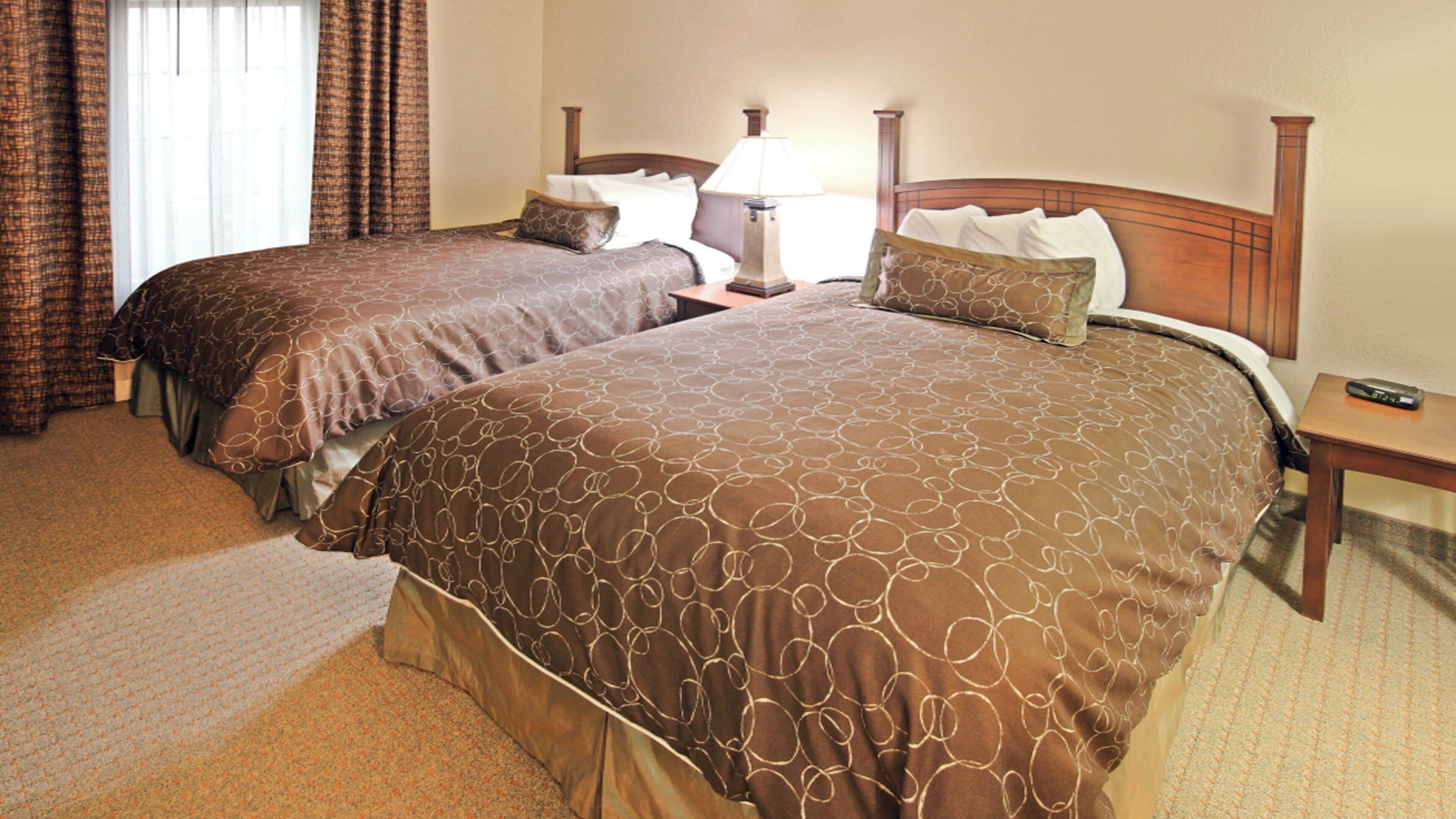 A hotel room with two beds, a nightstand, a lamp, and a remote control on the nightstand.