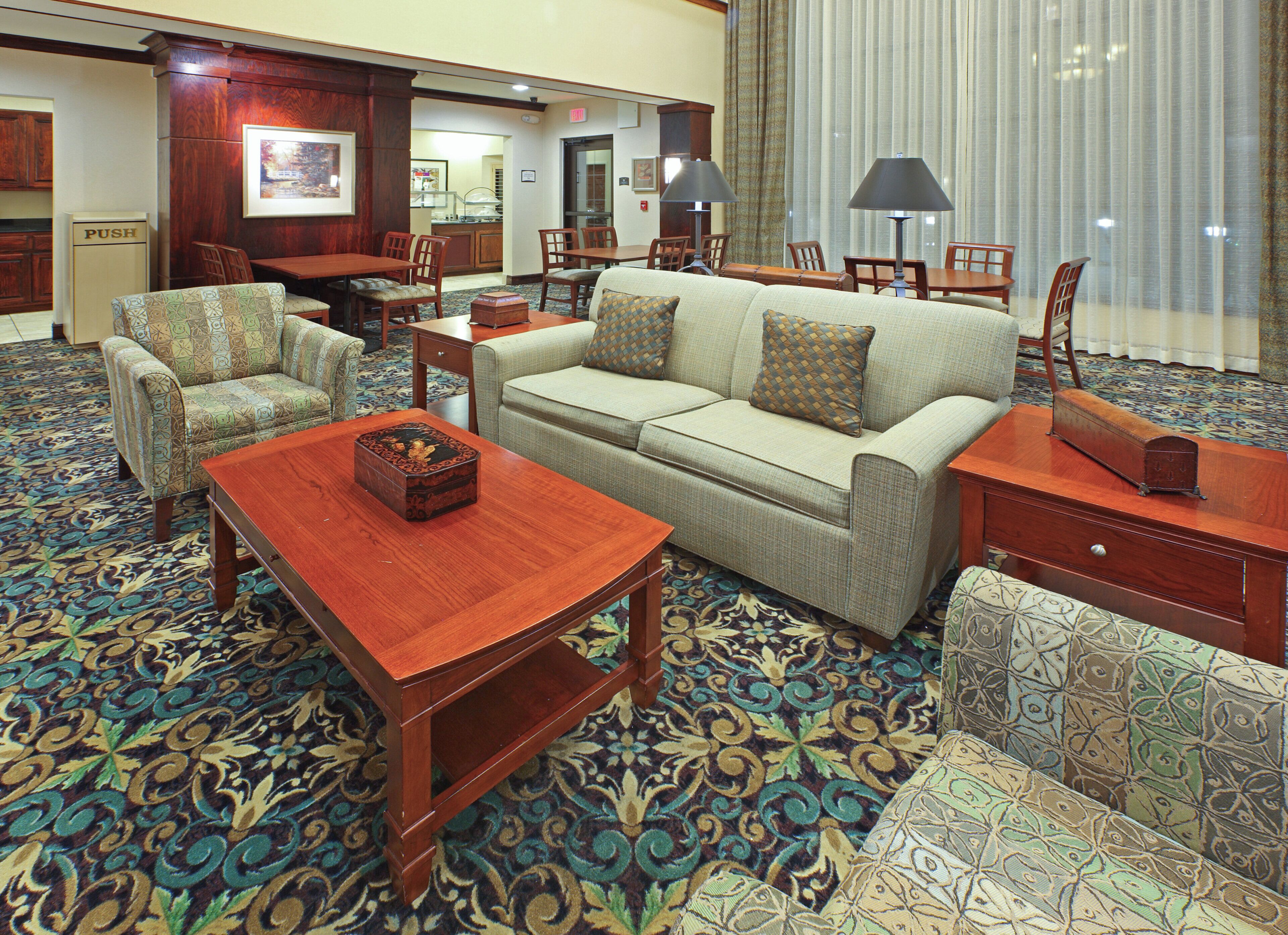 A hotel lobby with a patterned carpet, a sofa, armchairs, a wooden coffee table, and a dining area with a table and chairs.