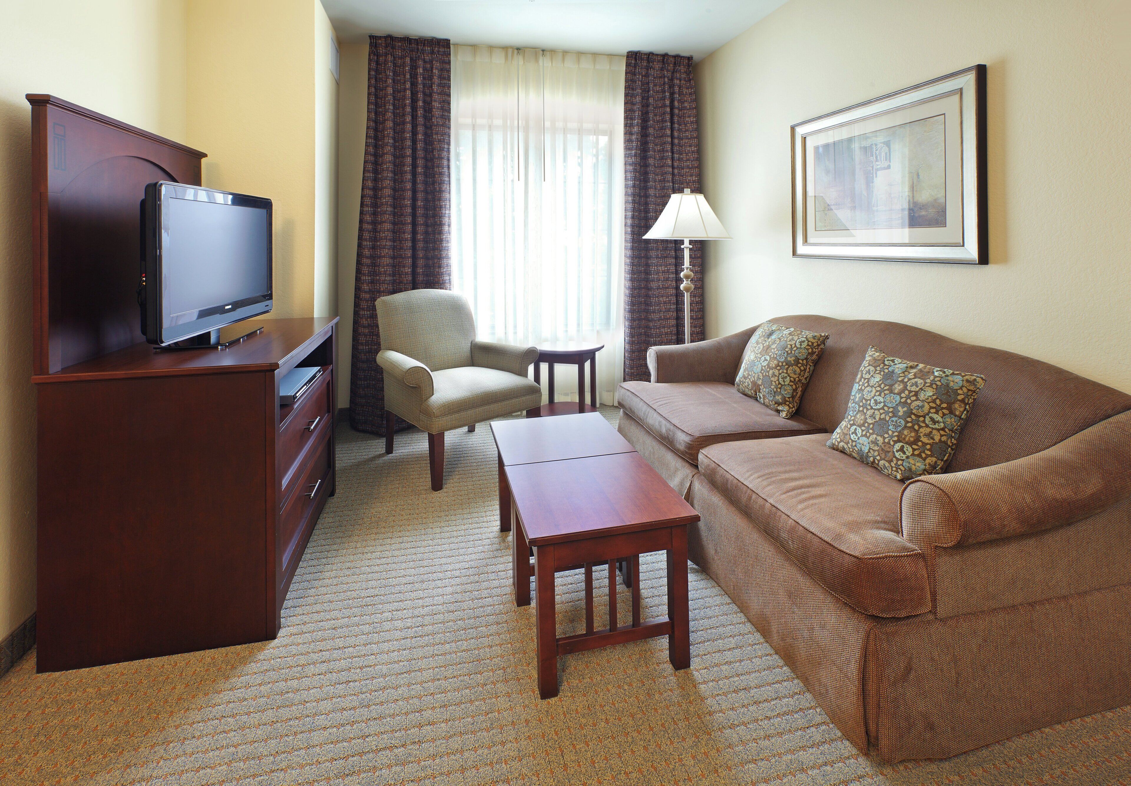A hotel room with a television, a sofa, a chair, a small table, and a lamp.