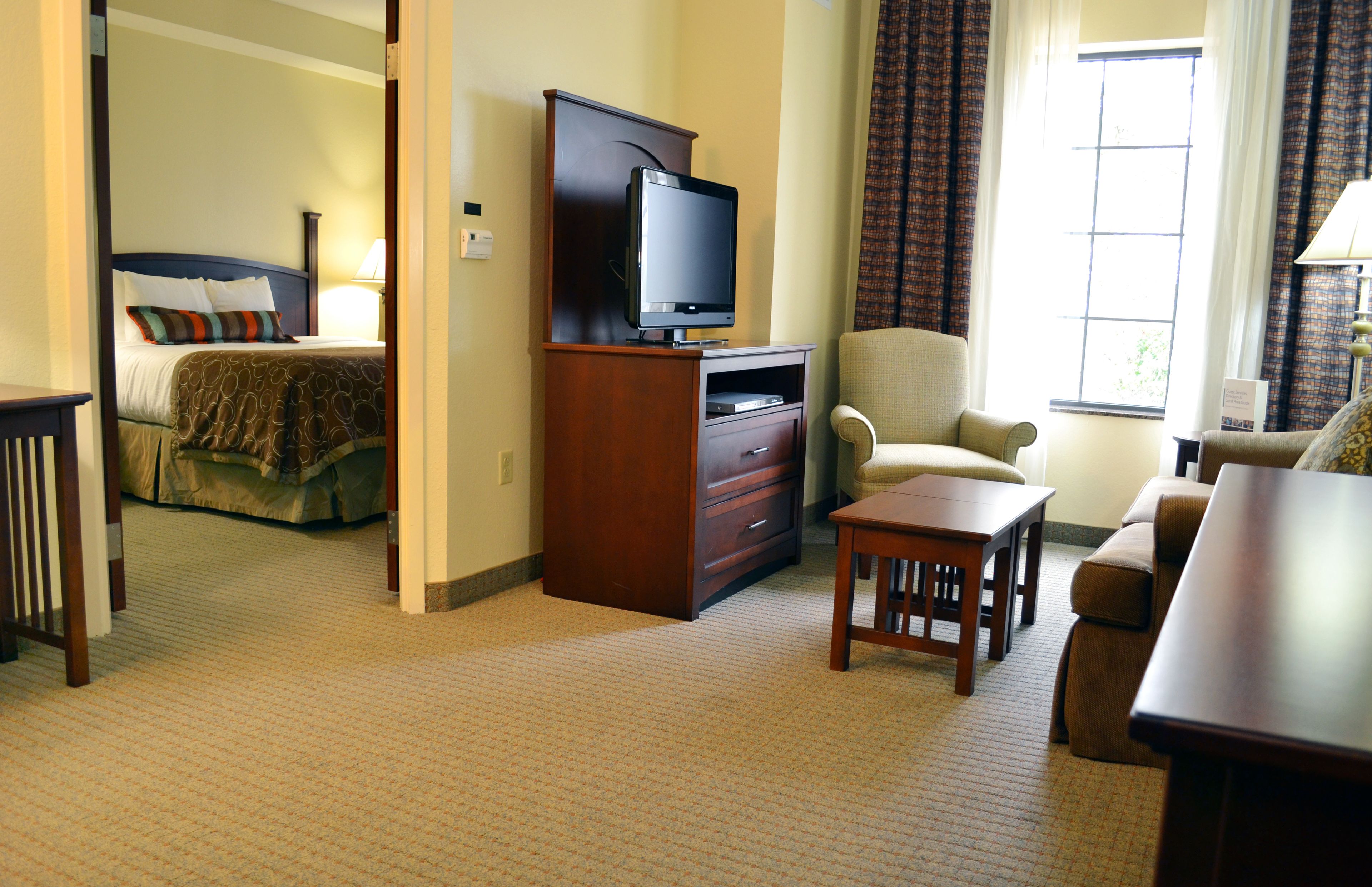 A hotel room with a bed, a television on a wooden stand, a sofa, a small table, and a window with curtains.