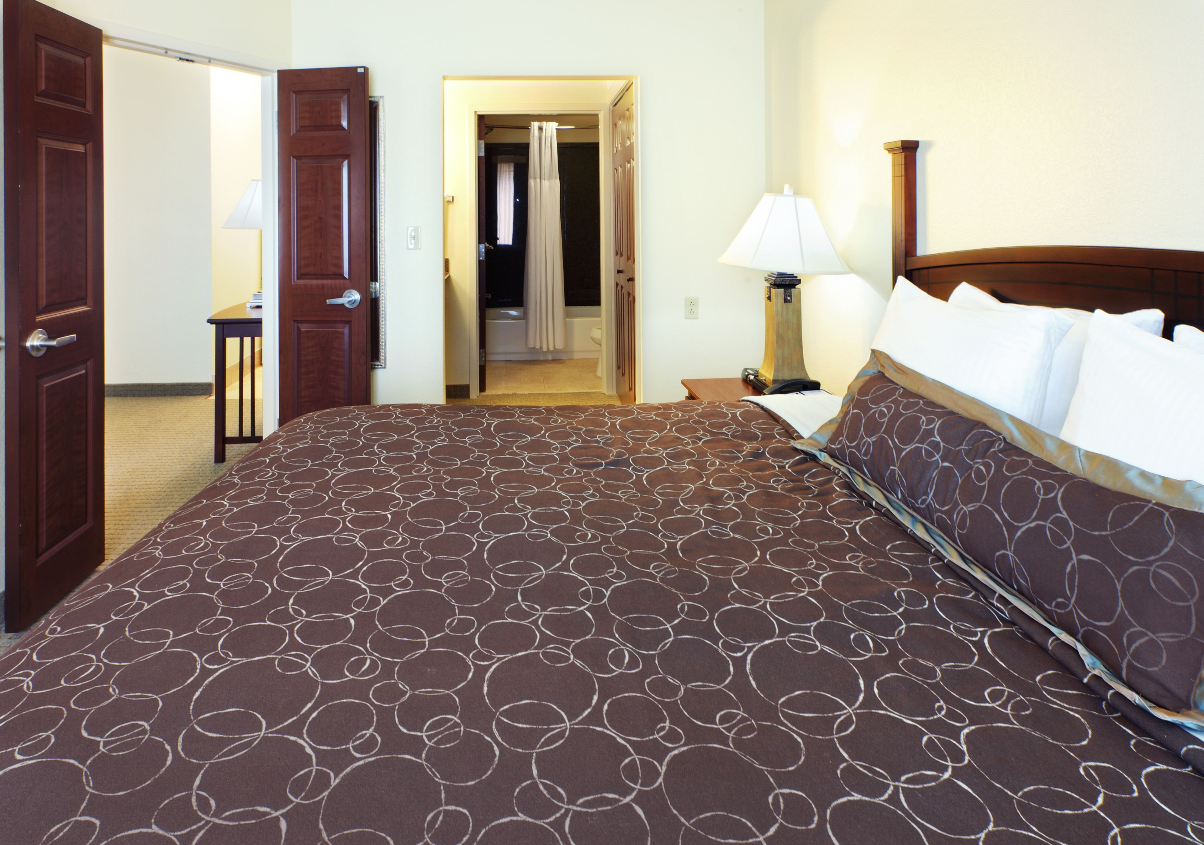Une chambre d’hôtel avec un grand lit, une table de chevet, une lampe et une salle de bain visible à travers une porte ouverte.