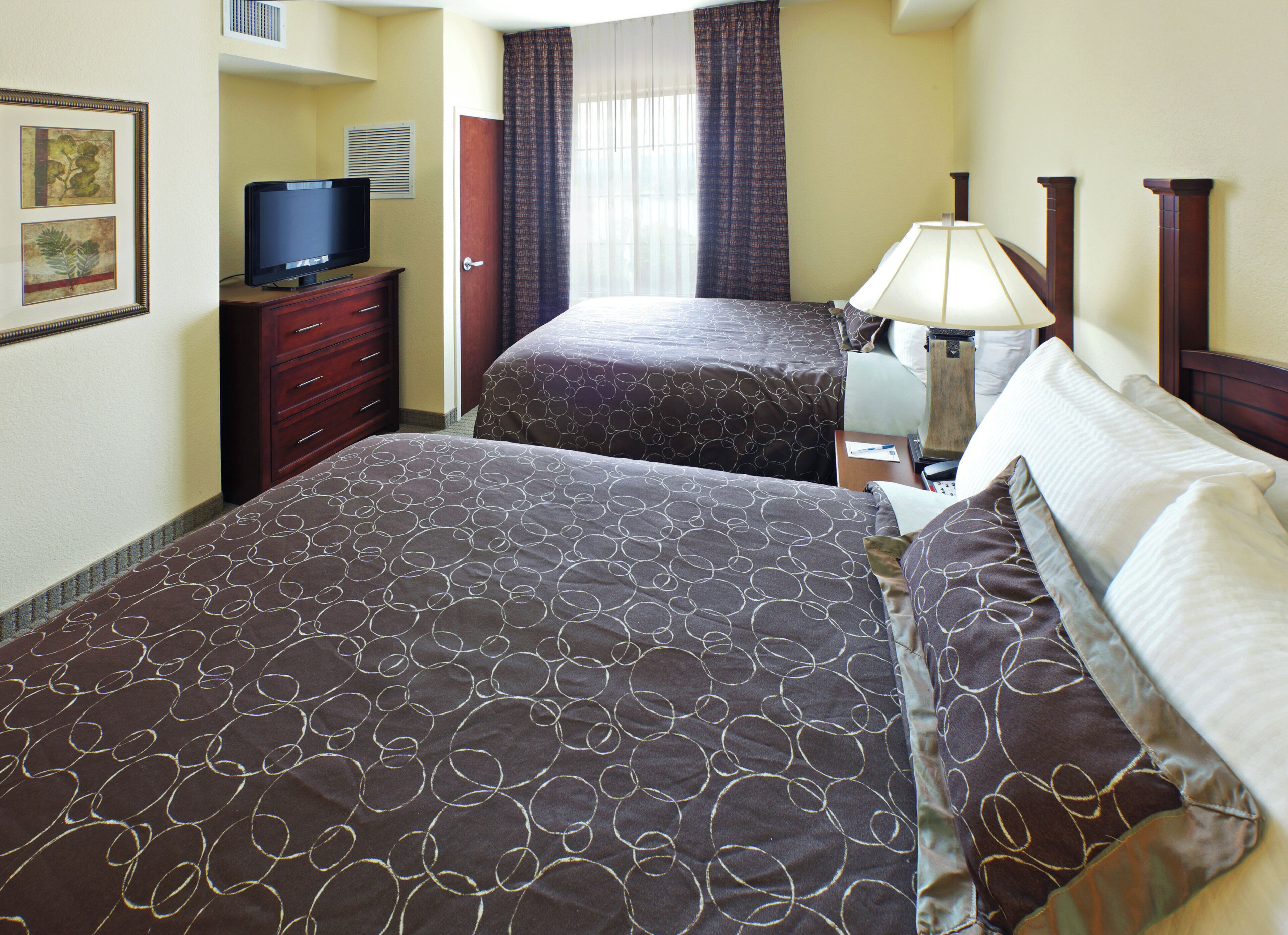 A hotel room with two beds, a television on a dresser, a lamp, and a window with curtains.