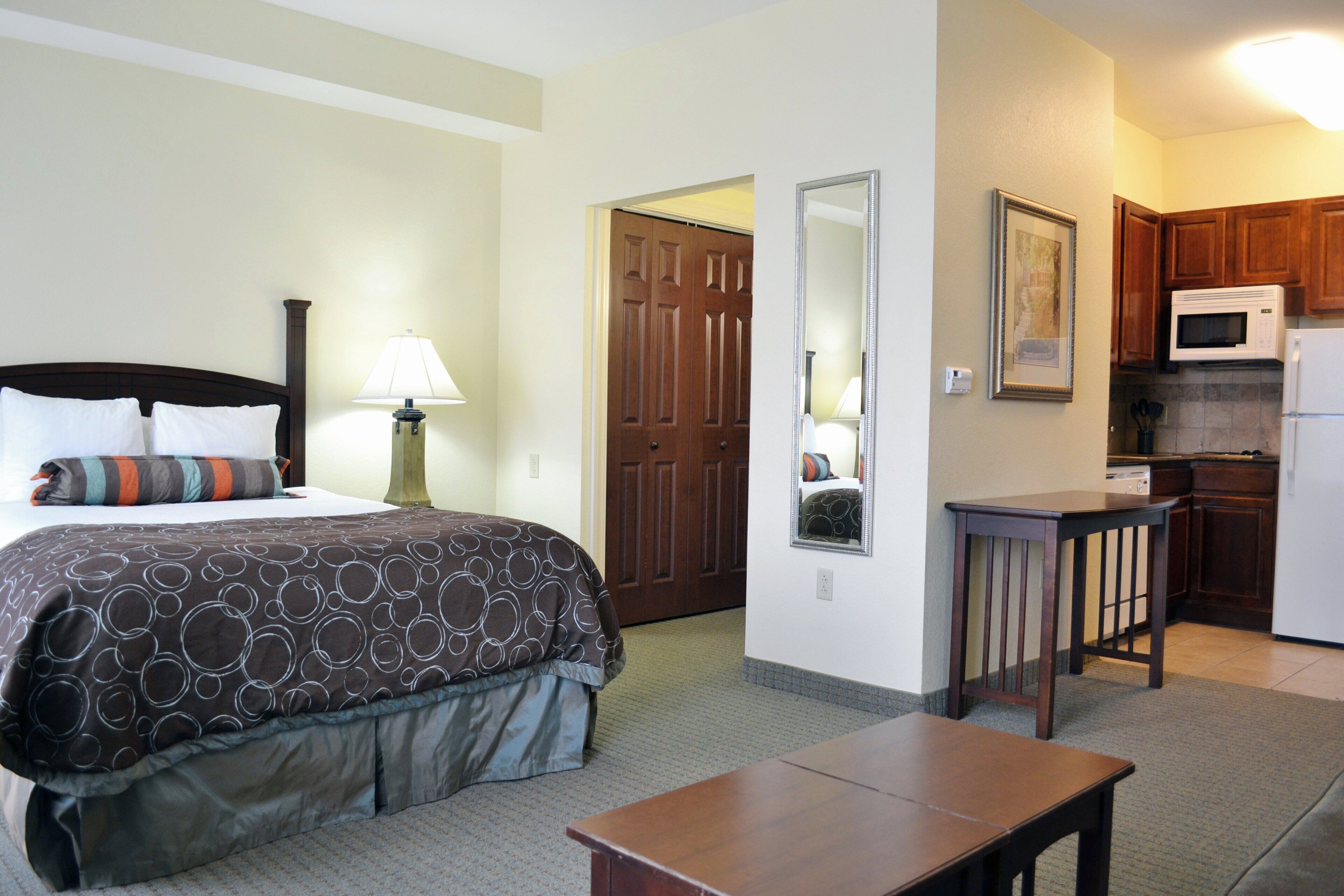 Una habitación de hotel con una cama, mesita de noche, lámpara y una kitchenette equipada con microondas y refrigerador.