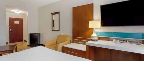 Una habitación de hotel con una cama, un escritorio, una silla, un televisor, una lámpara y un minibar.