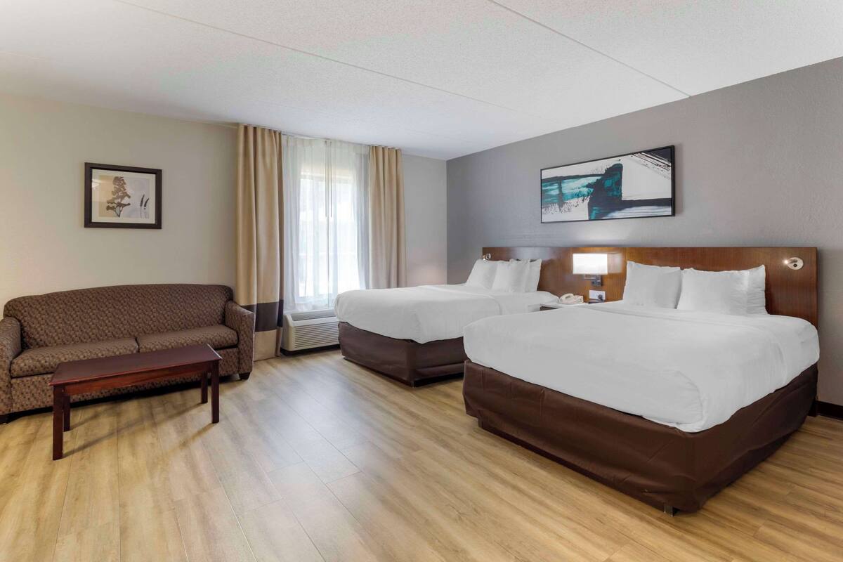 A hotel room with two beds, a sofa, a wooden floor, a window with curtains, and a framed picture on the wall.