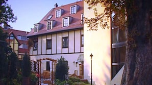 Un edificio de estilo europeo tradicional con tejado a dos aguas, numerosas ventanas y un pequeño jardín con una pérgola de madera.