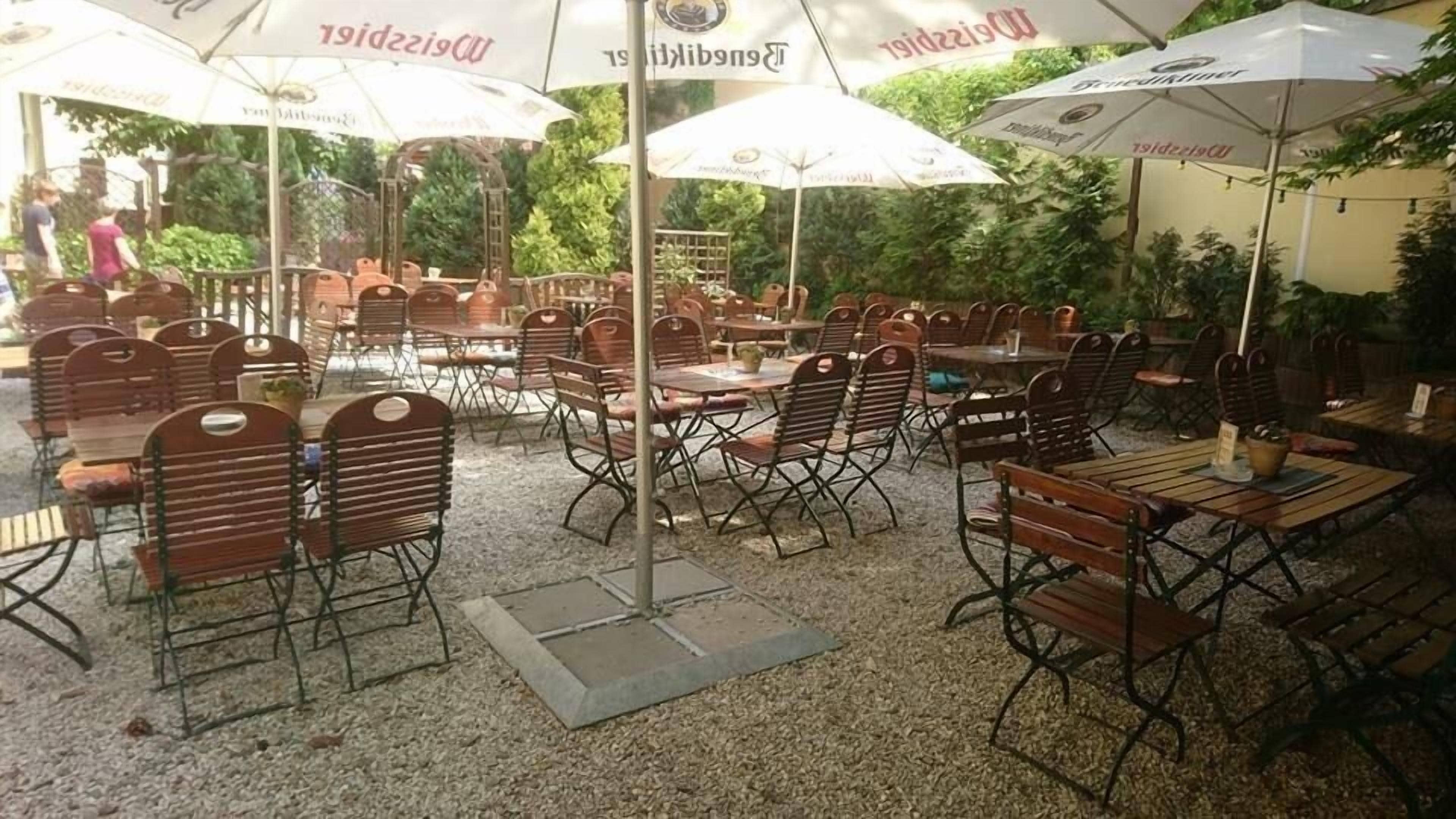 Un café al aire libre con mesas y sillas de madera, rodeado de sombrillas con texto visible.