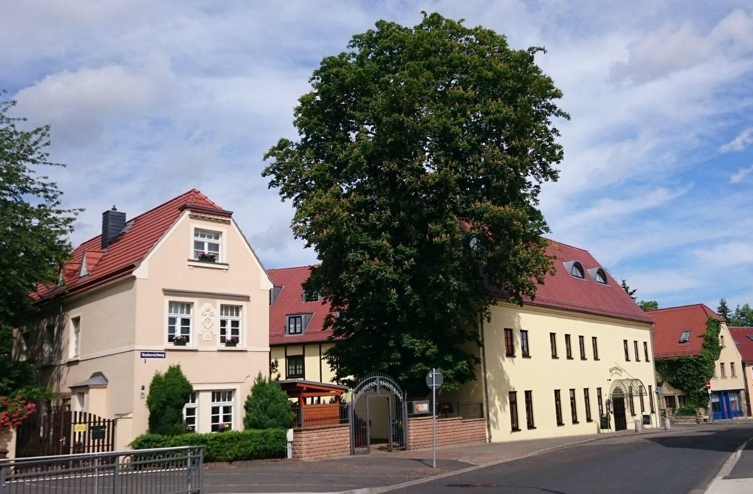 Un edificio tradicional europeo con tejado de tejas rojas, un gran árbol frente a él y una entrada con portones.