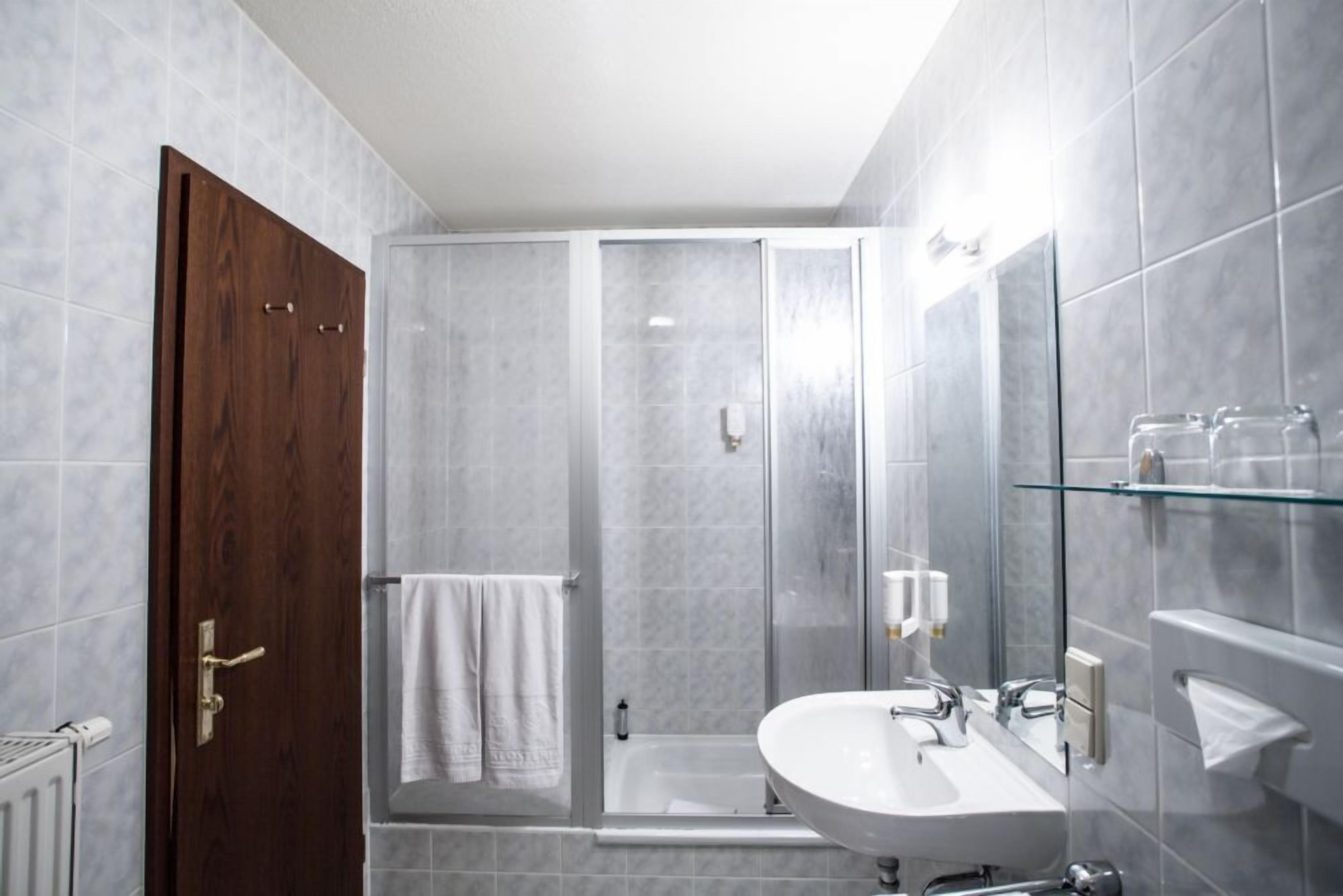 A modern bathroom with a wooden door, a glass shower enclosure, a white sink, and a glass shelf.