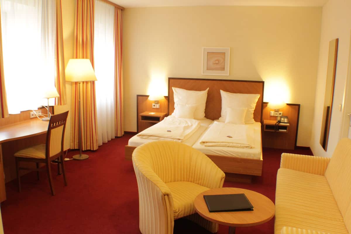 Una habitación de hotel con una cama, dos mesitas de noche con lámparas, un escritorio con una silla, un sofá y una pequeña mesa con un libro.