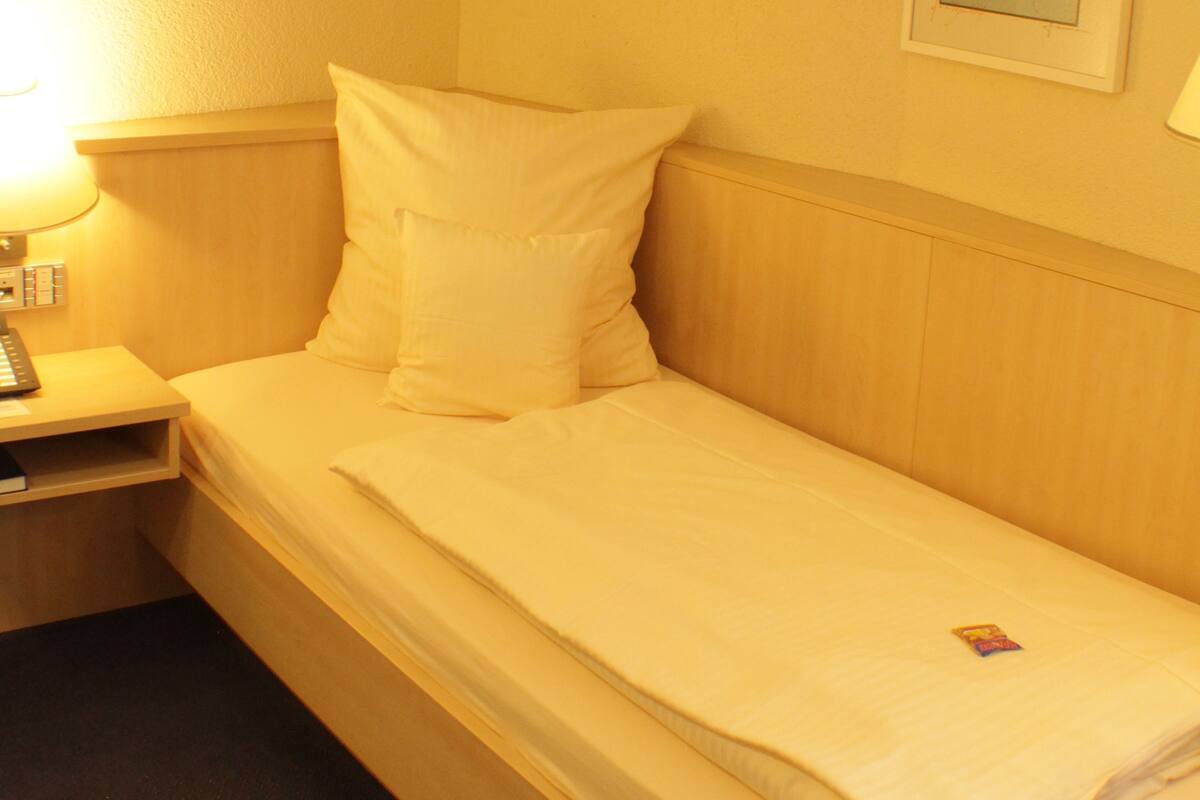 Una cama individual con ropa de cama blanca en una habitación con cabecera de madera y una mesita de noche pequeña con lámpara.