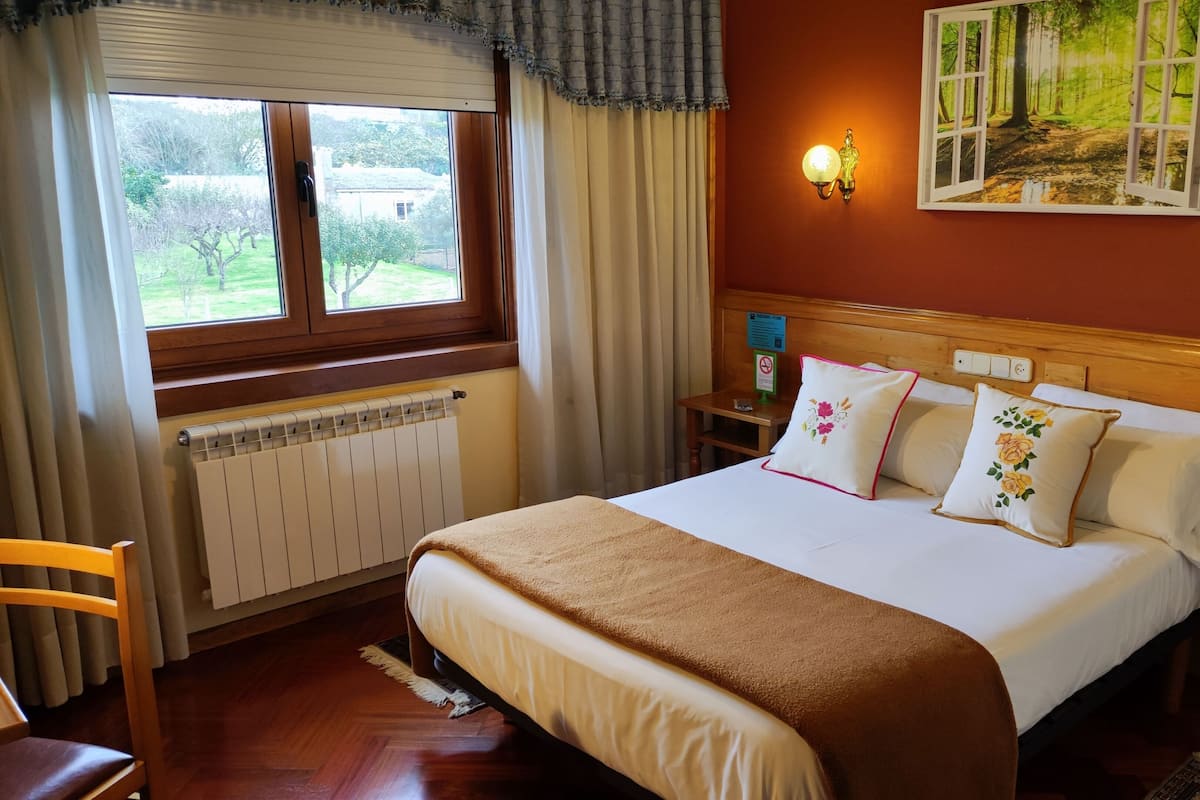 Quarto de hotel com uma cama grande, cabeceira de madeira, mesa de cabeceira com telefone, janela com cortinas, radiador e um quadro na parede.
