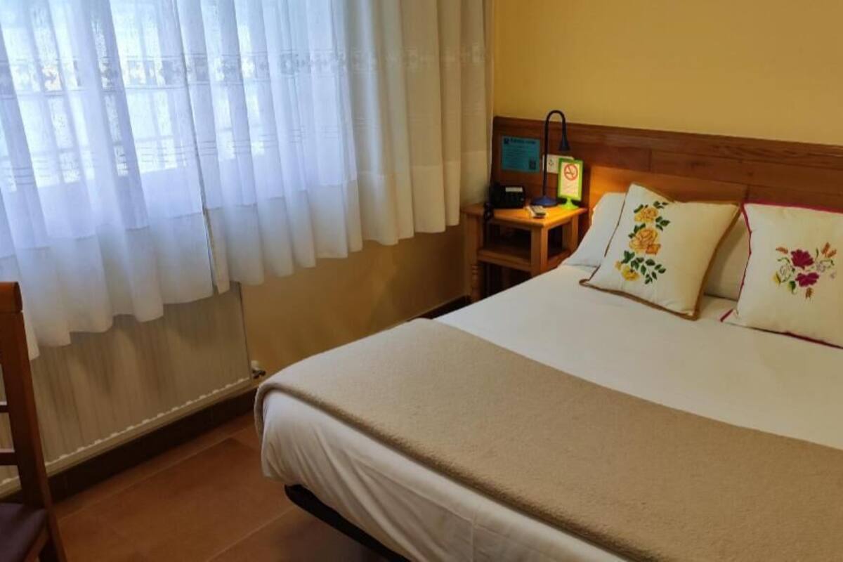 Quarto de hotel com cama, mesa de cabeceira, abajur e janela com cortinas.