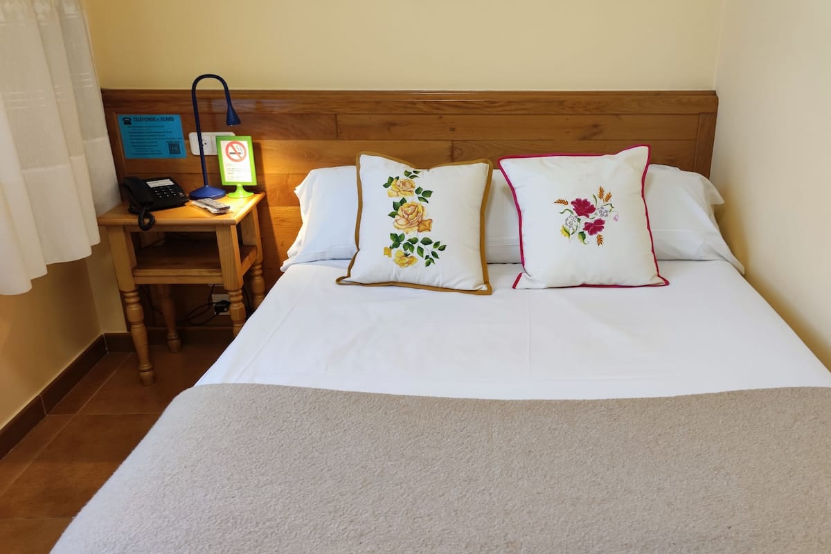 Quarto de hotel com cabeceira de madeira, duas almofadas bordadas, mesa de cabeceira com telefone e um letreiro, e uma luminária.