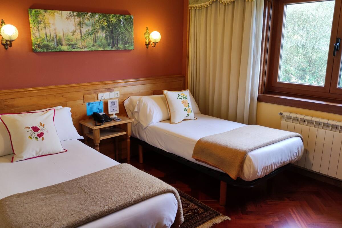 Quarto de hotel com duas camas, cabeceira de madeira, um quadro de uma floresta, telefone e janela com cortinas.