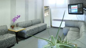 Un área de espera con sofás estampados, una mesita con planta y un televisor mostrando un anuncio.