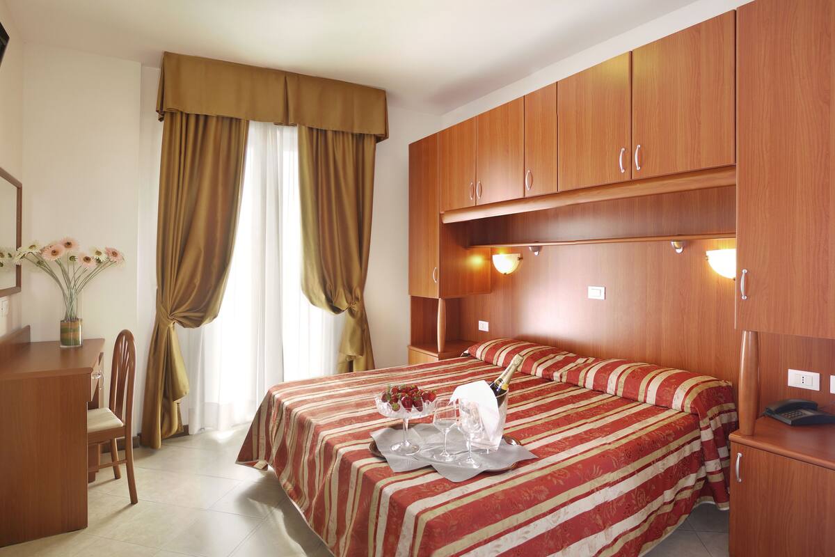 Habitación de hotel con cama, escritorio, silla, armario y ventana con cortinas.