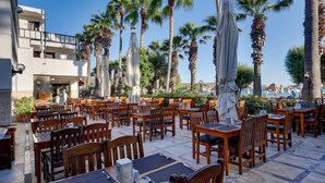 An outdoor dining area with wooden tables and chairs, surrounded by palm trees and a building with balconies. - Sami Beach Hotel (Bodrum)