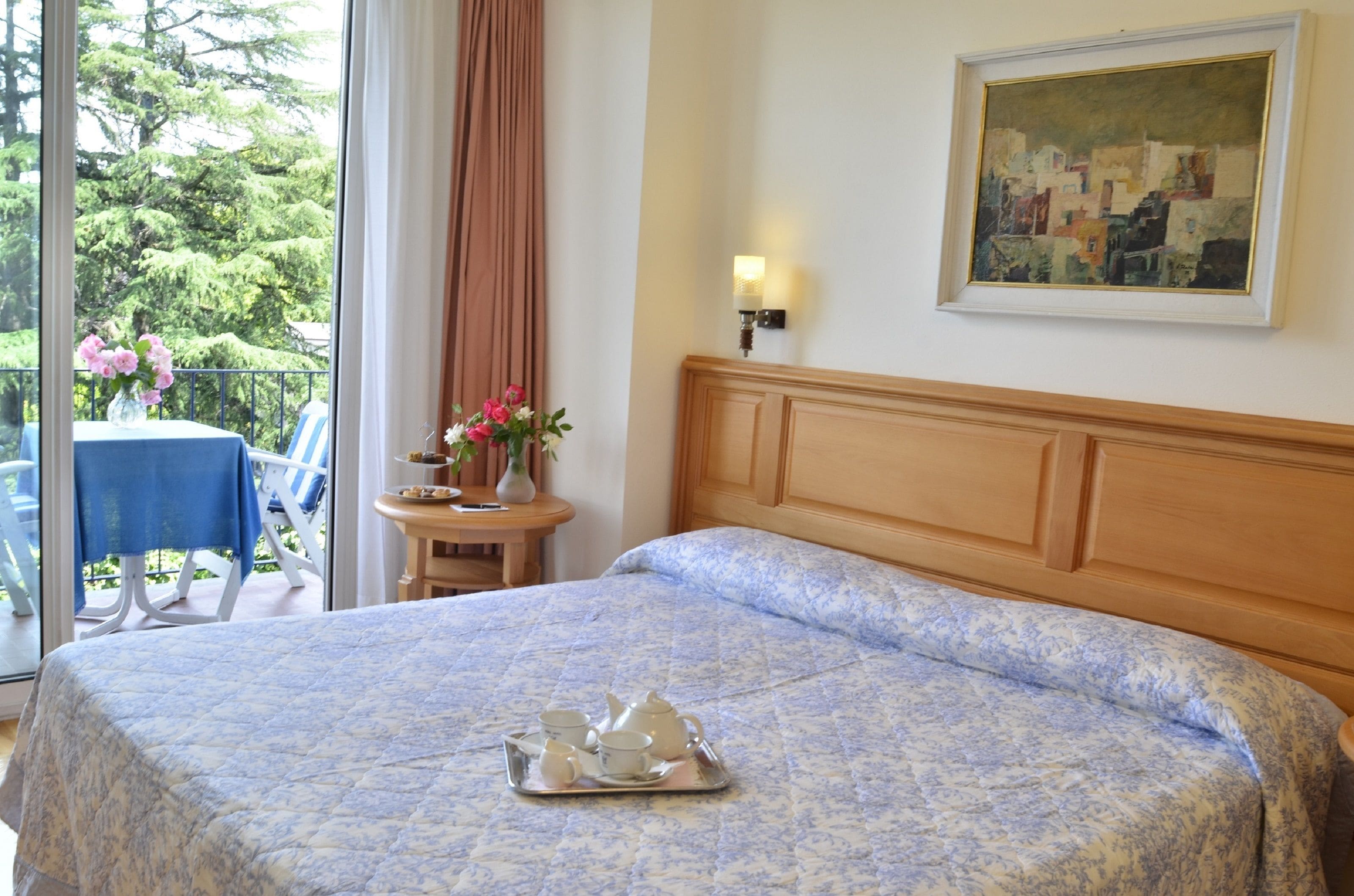 A hotel room with a bed, a painting, a table with a vase of flowers, and a balcony with a table and chairs.