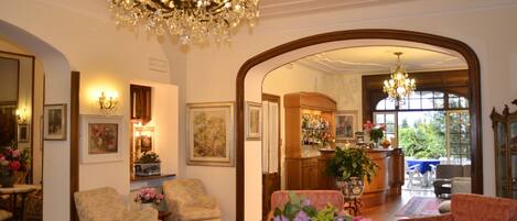 A spacious living room with a chandelier, floral arrangements, and a view of the garden through an archway.