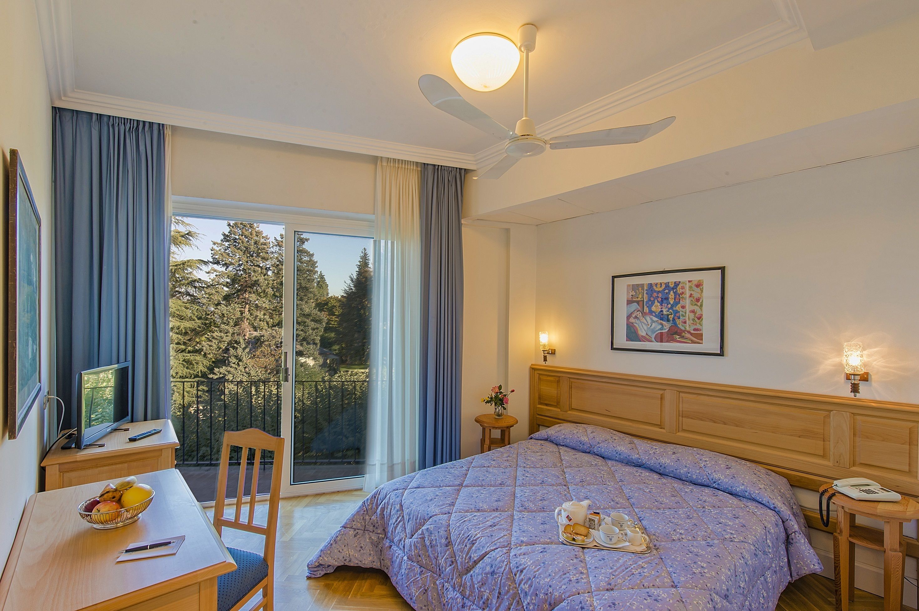Une chambre à coucher comprenant un lit, un bureau, une chaise, une table avec des fruits, un vase avec des fleurs, un tableau et un balcon avec vue sur les arbres.