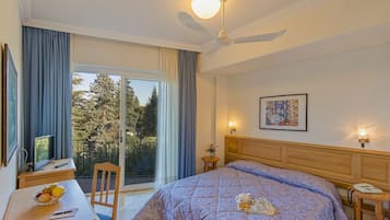 Une chambre à coucher comprenant un lit, un bureau, une chaise, une table avec des fruits, un vase avec des fleurs, un tableau et un balcon avec vue sur les arbres.