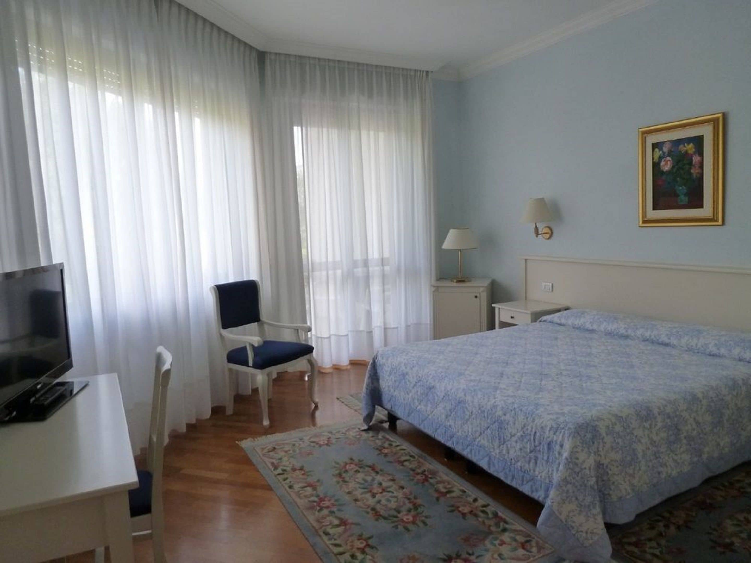 A hotel room with a bed, a desk with a computer, a chair, a lamp, and a painting on the wall.