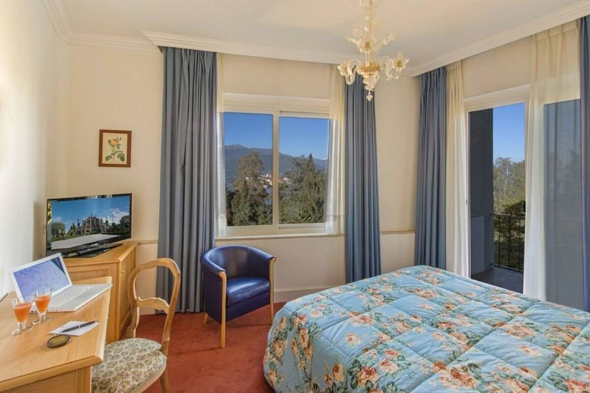 A hotel room with a bed, a desk with a laptop, a chair, a television, and a window with curtains.