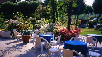 Terrassa exterior amb taules i cadires rodones, envoltada de verdura exuberant i plantes florejants.